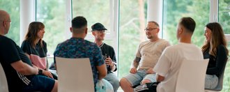 Eine diverse Gruppe von Menschen sitzt in einem Kreis auf weißen Stühlen und führt ein Gespräch in einer Gruppentherapie, in einem hellen Raum mit großen Fenstern.