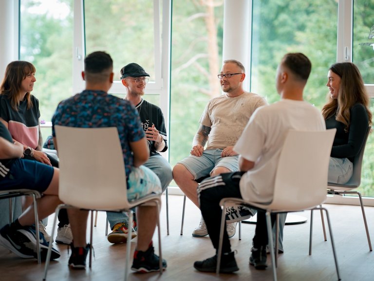 Eine diverse Gruppe von Menschen sitzt in einem Kreis auf weißen Stühlen und führt ein Gespräch in einer Gruppentherapie, in einem hellen Raum mit großen Fenstern.
