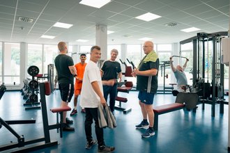 Eine Gruppe von Männern steht in einem hellen Fitnessstudio und unterhält sich. Im Vordergrund lächelt ein Mann in einem weißen T-Shirt in die Kamera. Im Hintergrund sind weitere Personen und Trainingsgeräte zu erkennen.