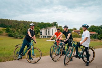 Vier junge Männer mit Fahrradhelm stehen mit ihren E-Bikes auf einem asphaltierten Weg und unterhalten sich. Im Hintergrund sind eine grüne Wiese, Bäume und ein weißes Gebäude zu sehen.