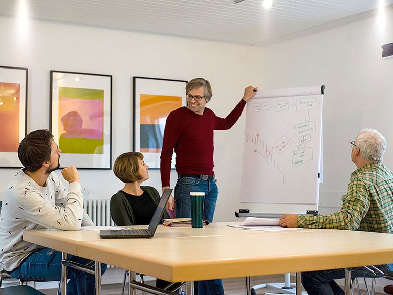 Besprechungsraum: ein junger Mann steht an einem Flipchart und erklärt einer kleinen Gruppe lächelnd sein "Gekritzel"