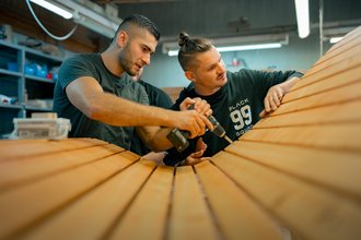 Zwei Männer arbeiten in einer Werkstatt an einer großen Holzkonstruktion. Einer bohrt mit einem Akkuschrauber in die Holzplanken, während der andere zusieht und hilft.