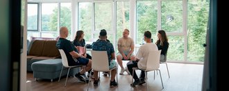 Sechs Personen sitzen in einem hellen Raum im Kreis und führen ein Gruppengespräch. Große Fenster im Hintergrund zeigen eine grüne Außenansicht.
