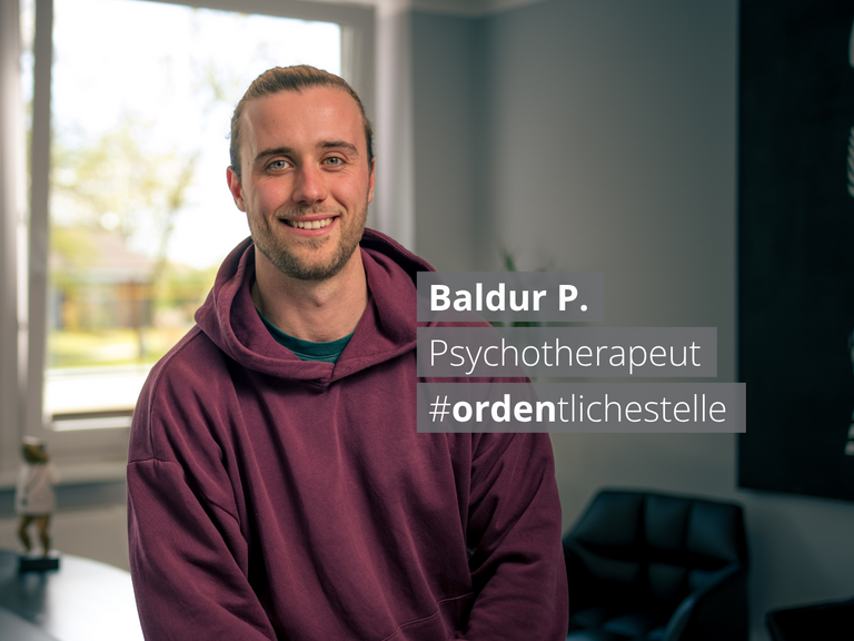 Ein Porträt von Baldur P., einem lächelnden Psychotherapeuten mit Bart in einem weinroten Hoodie. Er sitzt in einem hellen Büro. Ein Textfeld neben ihm nennt seinen Namen, Beruf und den Hashtag #ordentlichestelle.