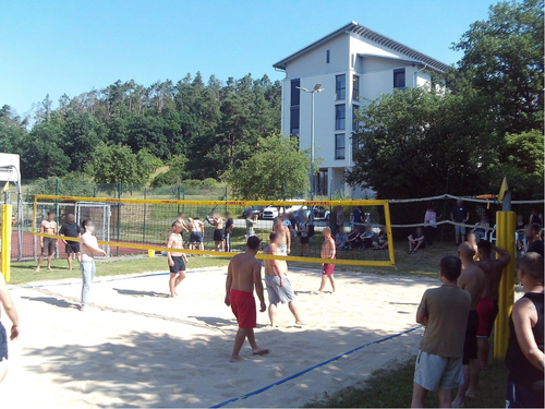 Eine Gruppe von Männern spielt Beachvolleyball auf einem Sandplatz an einem sonnigen Tag. Ein gelbes Netz teilt das Spielfeld. Im Hintergrund sind Bäume und ein weißes Gebäude zu sehen, während Zuschauer das Spiel beobachten.