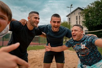 Eine Gruppe junger Männer in Sportkleidung umarmt sich herzlich auf einem Sandplatz. Sie lachen und wirken glücklich, als würden sie einen Sieg feiern. Im Hintergrund sind ein Netz und ein Gebäude zu sehen.