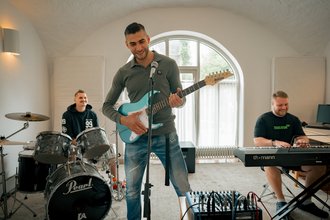 Ein Gitarrist und Sänger steht im Vordergrund und spielt eine hellblaue E-Gitarre. Im Hintergrund begleiten ihn links ein Schlagzeuger und rechts ein Keyboarder in einem hellen Probenraum.