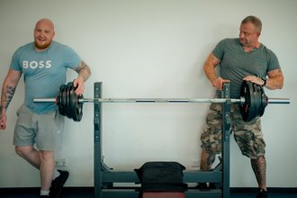 Zwei Männer stehen an einer Bankdrückbank und halten eine Langhantel mit Gewichten. Der Mann links im blauen T-Shirt lächelt, während der Mann rechts im grauen Shirt die Gewichte auf der Hantel betrachtet.