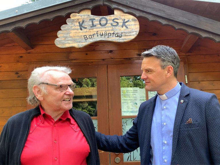 Pater Jörg vor dem Kiosk "Barfußpfad" hört einem älteren Mann zu