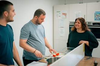 Drei Menschen in einer Küche. Ein Mann in der Mitte schneidet Gemüse auf einem Brett. Eine Frau rechts lächelt und beobachtet ihn, während ein weiterer Mann links daneben steht und zuschaut.