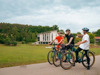 Drei Radfahrer stehen auf einem asphaltierten Weg und unterhalten sich. Einer zeigt auf ein modernes weißes Haus im Hintergrund, das umgeben von einer grünen Wiese und Bäumen ist.