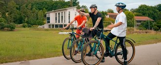 Drei Radfahrer stehen auf einem asphaltierten Weg und unterhalten sich. Einer zeigt auf ein modernes weißes Haus im Hintergrund, das umgeben von einer grünen Wiese und Bäumen ist.