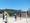 Vier Personen spielen Beachvolleyball auf einem Sandplatz. Ein Spieler im blauen Shirt schlägt den Ball über das Netz. Im Hintergrund sind Bäume, ein weißes Haus und ein Tennisplatz unter einem klaren blauen Himmel zu sehen.