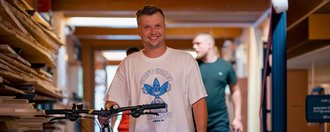 Ein lächelnder Mann in einem weißen T-Shirt steht mit einem Fahrrad in einer Werkstatt. Im Hintergrund sind Holzstapel und ein weiterer Mann zu sehen.