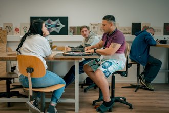 Vier Personen arbeiten in einem hellen Raum mit Kunstwerken im Hintergrund. Im Vordergrund sitzen eine Frau und ein Mann an einem Tisch, wobei der Mann an einem Laptop arbeitet. Zwei weitere Personen sind im Hintergrund an ihren Plätzen beschäftigt.