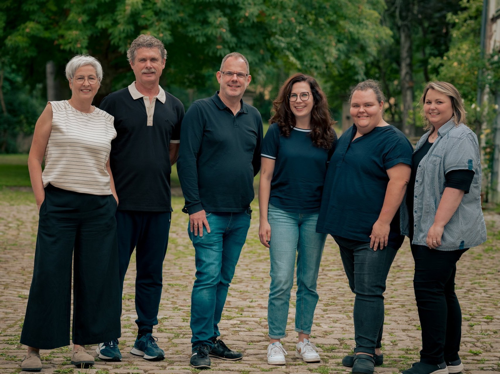 Gruppenfoto von sechs Personen, die nebeneinander im Freien stehen und in die Kamera lächeln. Im Hintergrund sind Bäume und ein Haus zu erkennen.