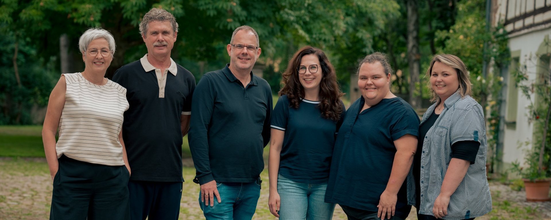 Gruppenfoto von sechs Personen, die nebeneinander im Freien stehen und in die Kamera lächeln. Im Hintergrund sind Bäume und ein Haus zu erkennen.