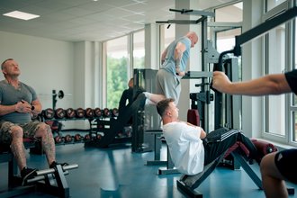 In einem hellen Fitnessstudio trainieren mehrere Männer. Ein Mann ruht auf einer Bank, ein anderer führt Beinheben aus, und im Hintergrund hängt ein Mann an einer Stange. Verschiedene Fitnessgeräte und Hanteln sind im Raum verteilt.