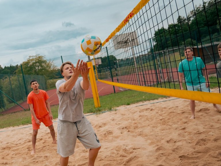 Männer spielen Beachvolleyball auf einem Sandplatz. Ein Spieler in der Mitte stößt den Ball über das Netz, während seine Mitspieler und Gegner das Spiel verfolgen.