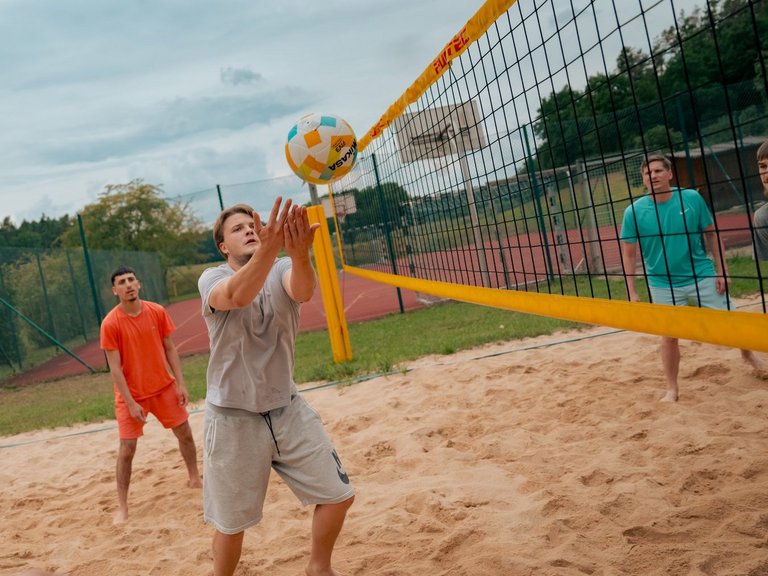 Männer spielen Beachvolleyball auf einem Sandplatz. Ein Spieler in der Mitte stößt den Ball über das Netz, während seine Mitspieler und Gegner das Spiel verfolgen.