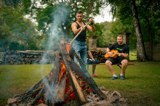Zwei Männer verbringen Zeit im Freien bei einem Lagerfeuer. Ein Mann steht und hantiert mit einem Stock am brennenden Holz, während der andere auf einer Bank sitzt und Gitarre spielt.