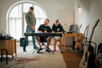 Drei Männer in einem hellen Musikraum. Zwei sitzen an einem Synthesizer und spielen, während ein dritter Mann links daneben steht. Im Vordergrund stehen mehrere Gitarren auf Ständern, und ein großes Bogenfenster befindet sich im Hintergrund.
