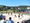 Beachvolleyball-Spieler kämpfen um den Ball auf einem Sandplatz unter blauem Himmel. Ein gelbes Netz teilt das Feld. Zuschauer verfolgen das Spiel im Vordergrund, während im Hintergrund Bäume und ein Basketballkorb stehen.