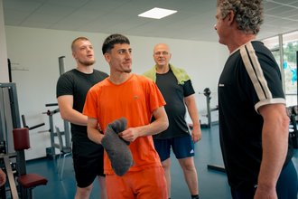 Vier Männer in Sportkleidung stehen in einem Fitnessstudio. Ein Mann rechts im Bild spricht zu den anderen drei. Der Mann in der Mitte trägt ein oranges T-Shirt und hält ein graues Handtuch. Im Hintergrund sind Fitnessgeräte zu sehen.
