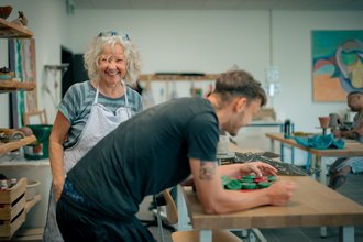 Eine fröhliche Frau mit grauen Locken und Schürze steht in einer Werkstatt und lächelt in die Kamera. Im Vordergrund arbeitet ein Mann konzentriert an einem Tisch mit grünem Material.