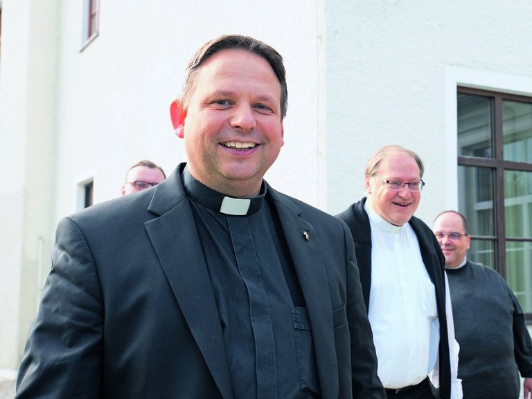 Vier Männer gehen vor einem weißen Gebäude. Der Mann links im Vordergrund trägt einen schwarzen Anzug mit Priesterkragen und lächelt. Rechts neben ihm geht ein Mann mit Brille und schwarzem Hemd. Im Hintergrund sind zwei weitere Männer, einer davon in weißer Kleidung.