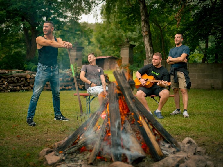 Vier Männer versammeln sich um ein Lagerfeuer im Freien. Einer spielt Gitarre, während die anderen beiden entspannt zuschauen.