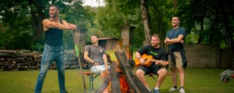 Vier Männer versammeln sich um ein Lagerfeuer im Freien. Einer spielt Gitarre, während die anderen beiden entspannt zuschauen.