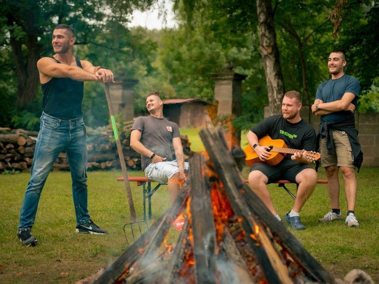 Vier Männer versammeln sich um ein Lagerfeuer im Freien. Einer spielt Gitarre, während die anderen beiden entspannt zuschauen.